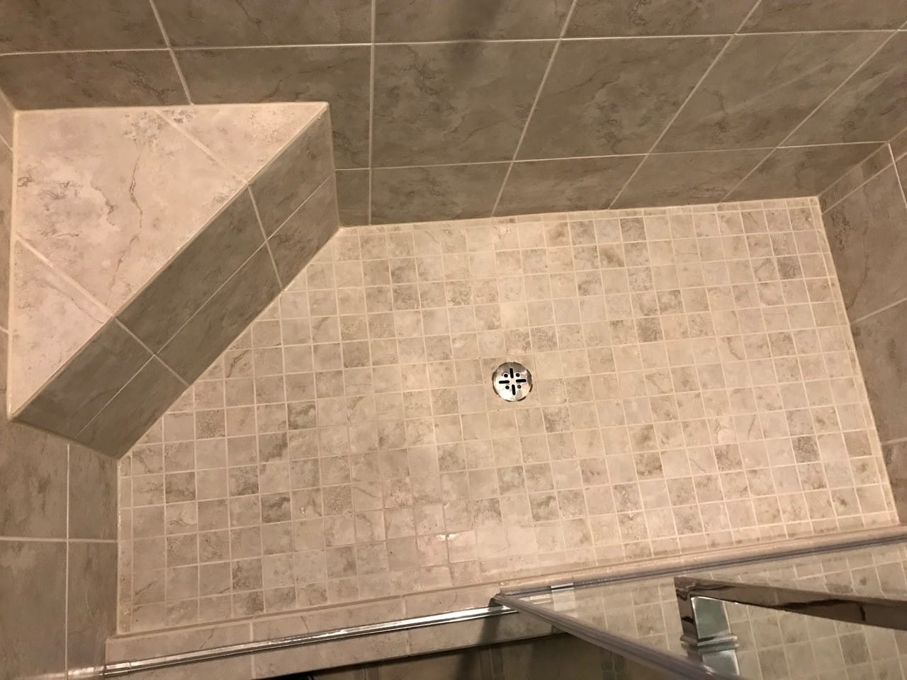 Mosaic-tiled shower floor sloped to a centre drain, with a tiled corner bench and stone-look wall tile