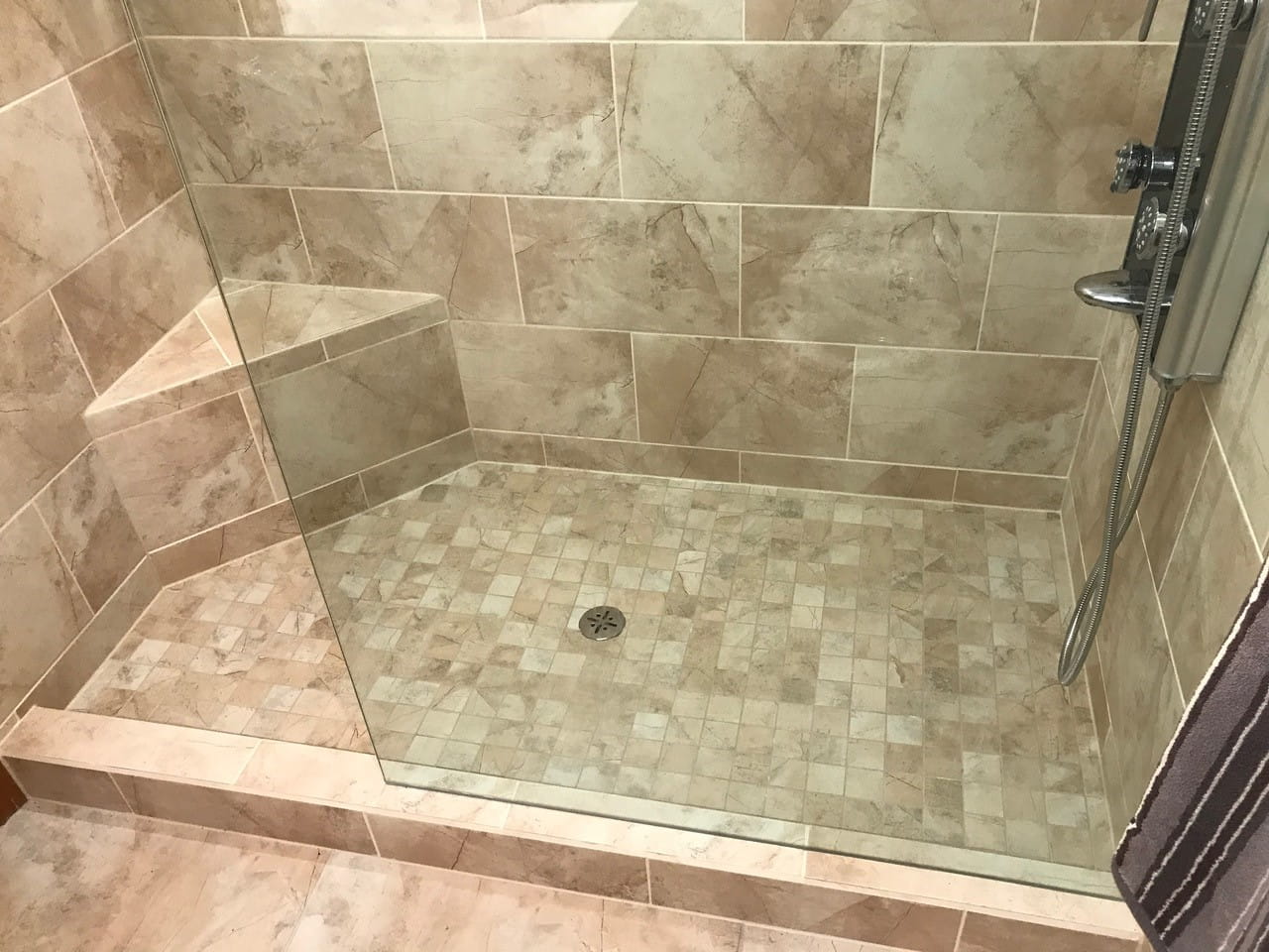 Large walk-in shower with beige stone tile, corner seat and mosaic floor